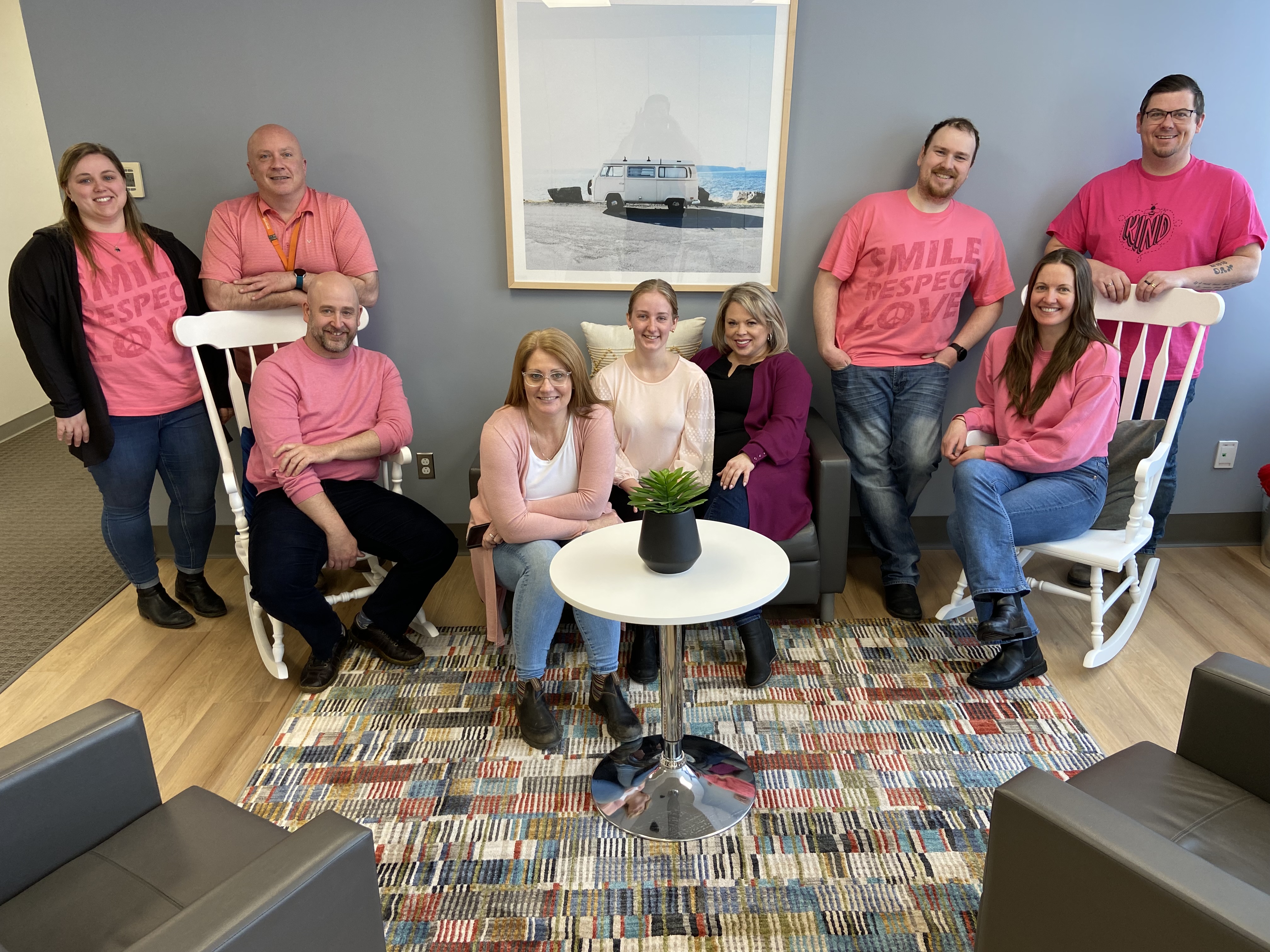 Office staff sitting on a variety of seating, smiling and wearing pink shirts.
