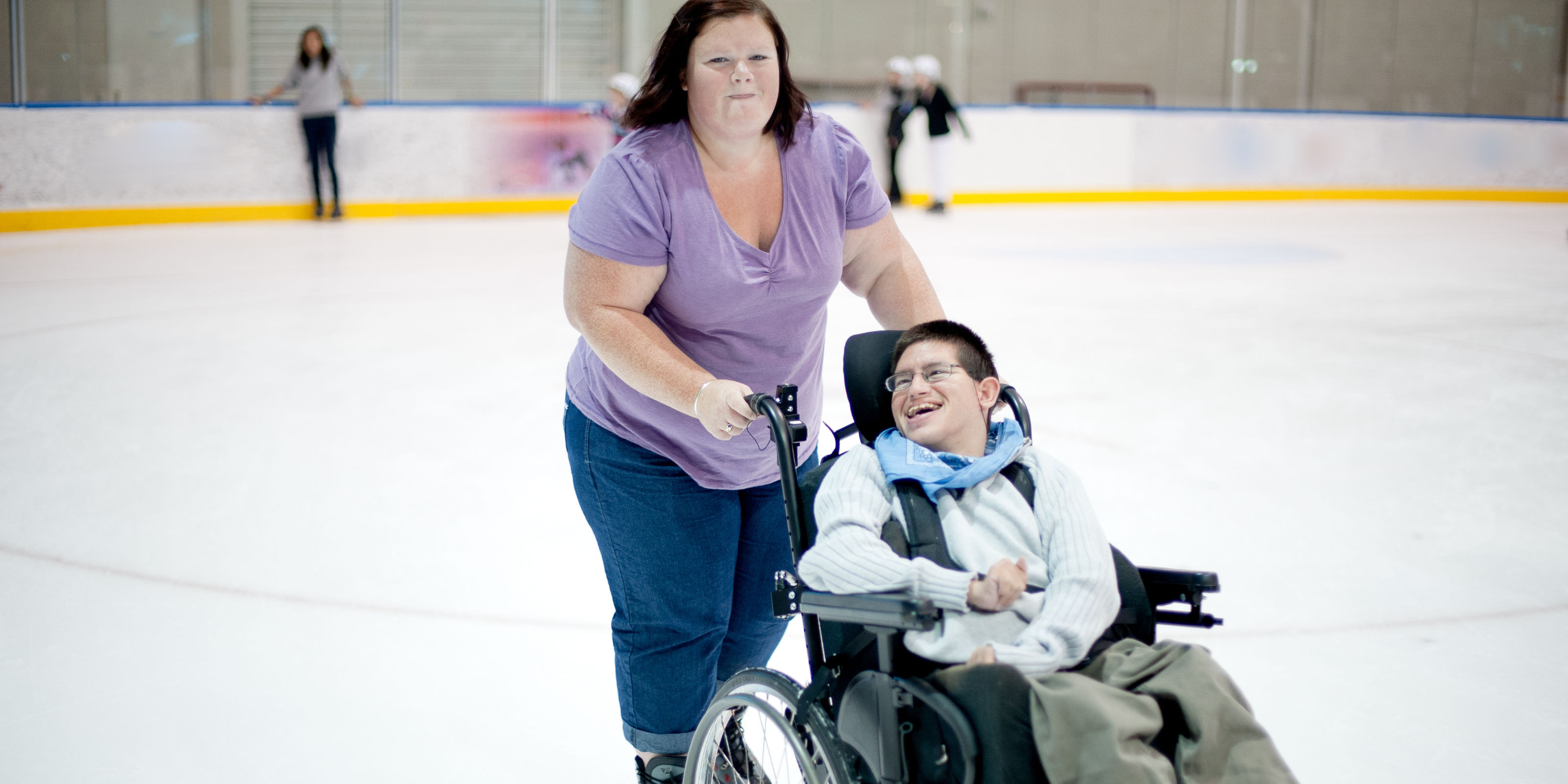 A carer pushes the wheelchair of a client across a skating rink.