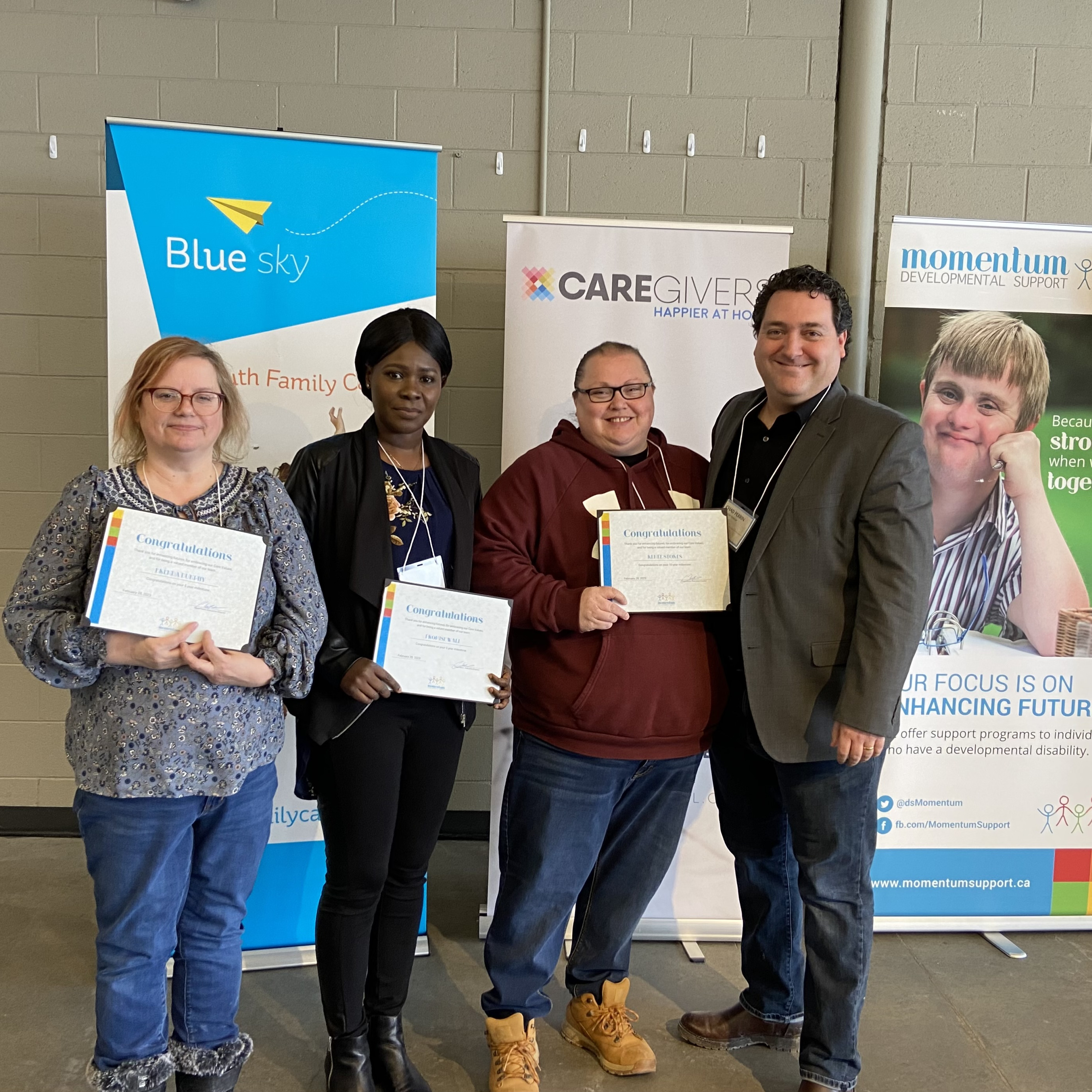 A group of four people hold up certifications in front of signage for Caregivers, Momentum, and Blue Sky.